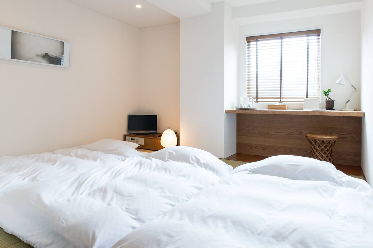 A bedroom with a bed, a desk, a computer, and a window with blinds.