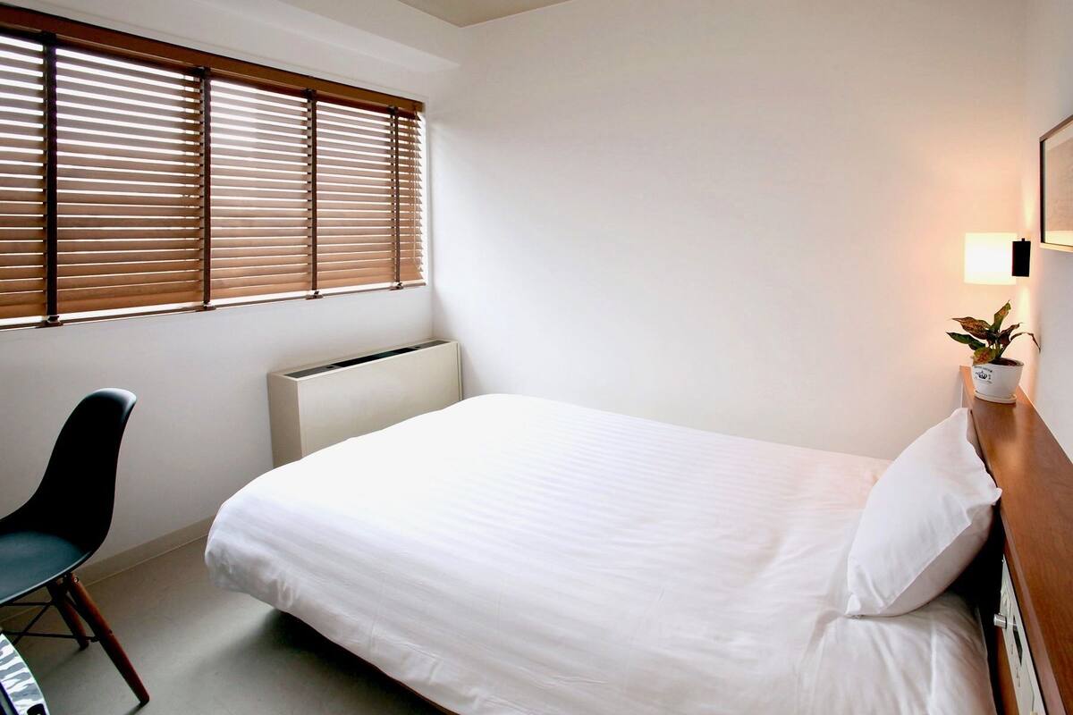 A neatly made bed in a hotel room with a radiator, a chair, and a potted plant on a bedside table.
