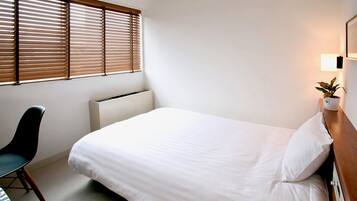 A neatly made bed in a hotel room with a radiator, a chair, and a potted plant on a bedside table.