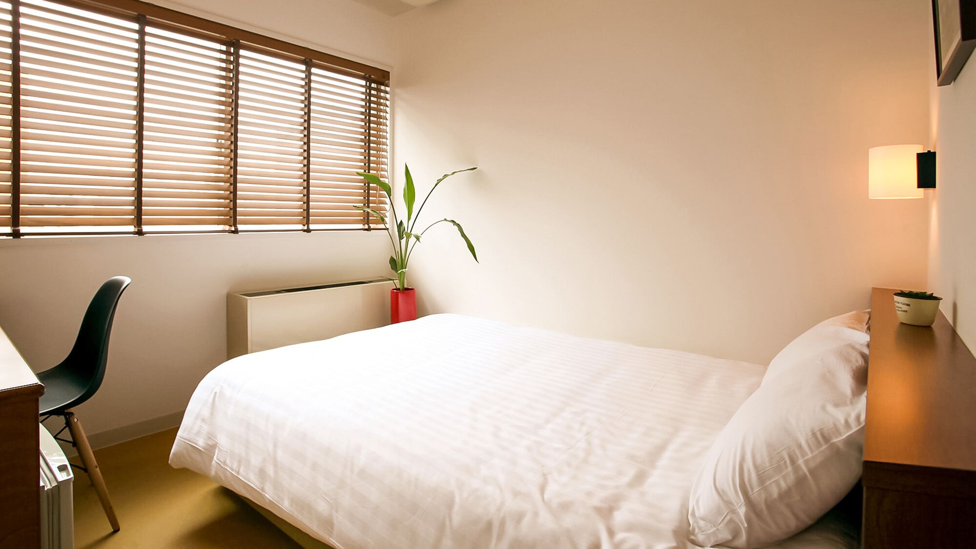 Une chambre à coucher comprenant un lit, un bureau, une chaise, un radiateur, une plante en pot et une lampe fixée au mur.