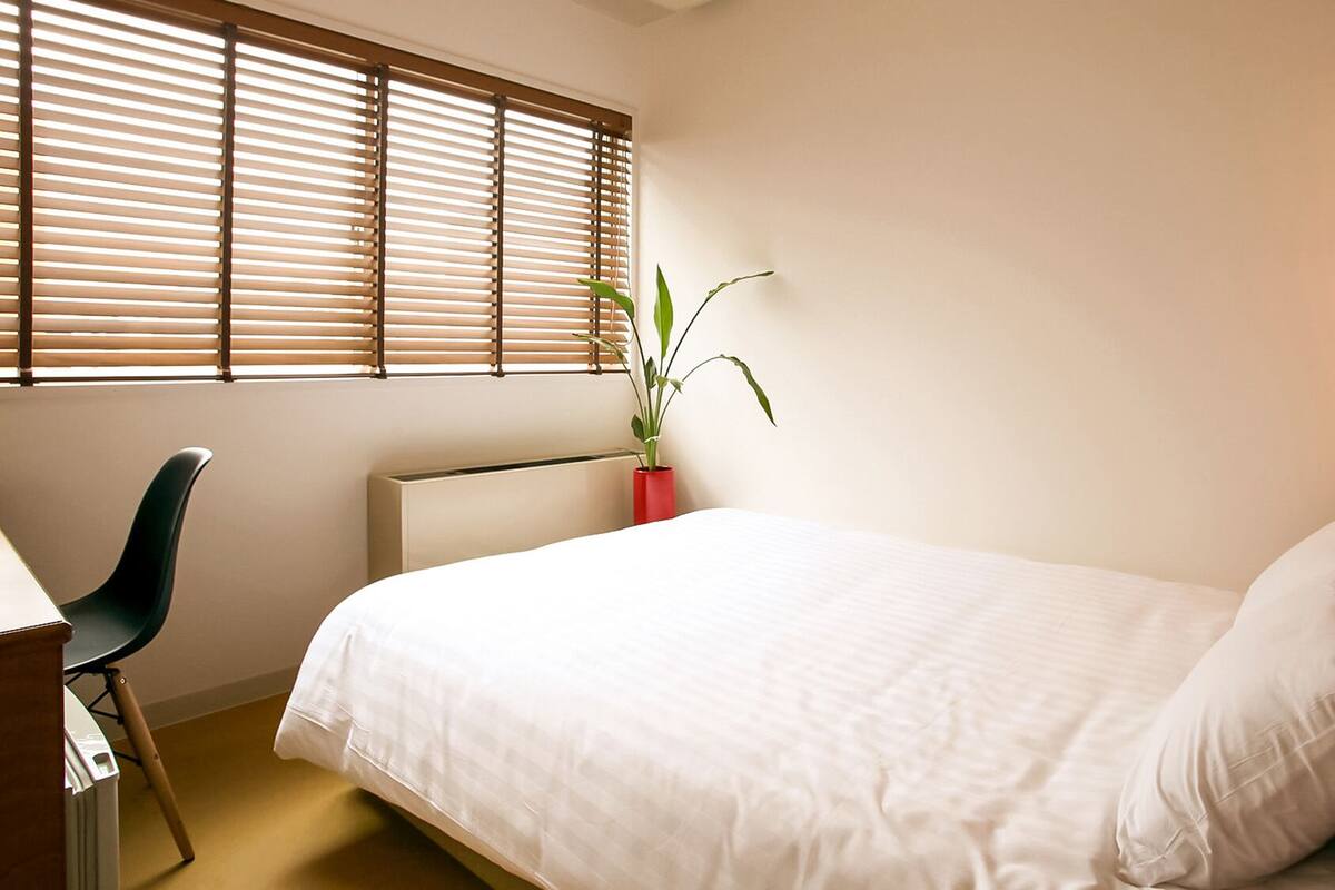 A bedroom with a bed, a desk, a chair, a radiator, a potted plant, and a wall-mounted lamp.