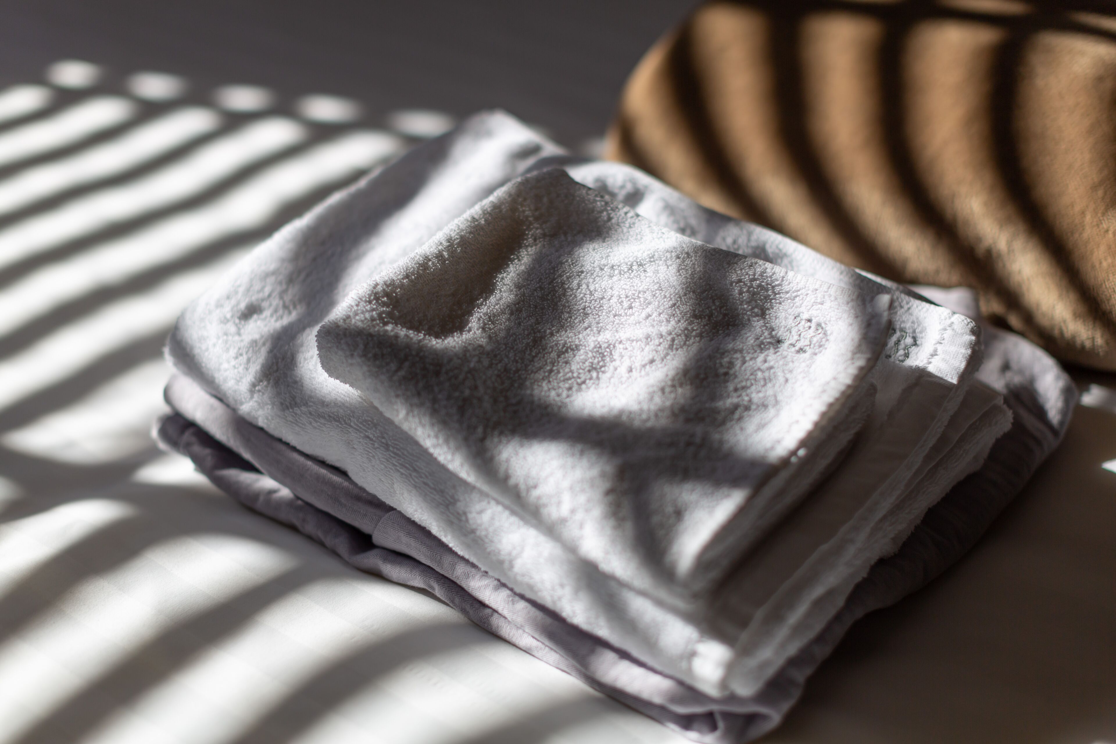 A neatly folded towel on a bed with a patterned pillow and a striped shadow cast on the wall.