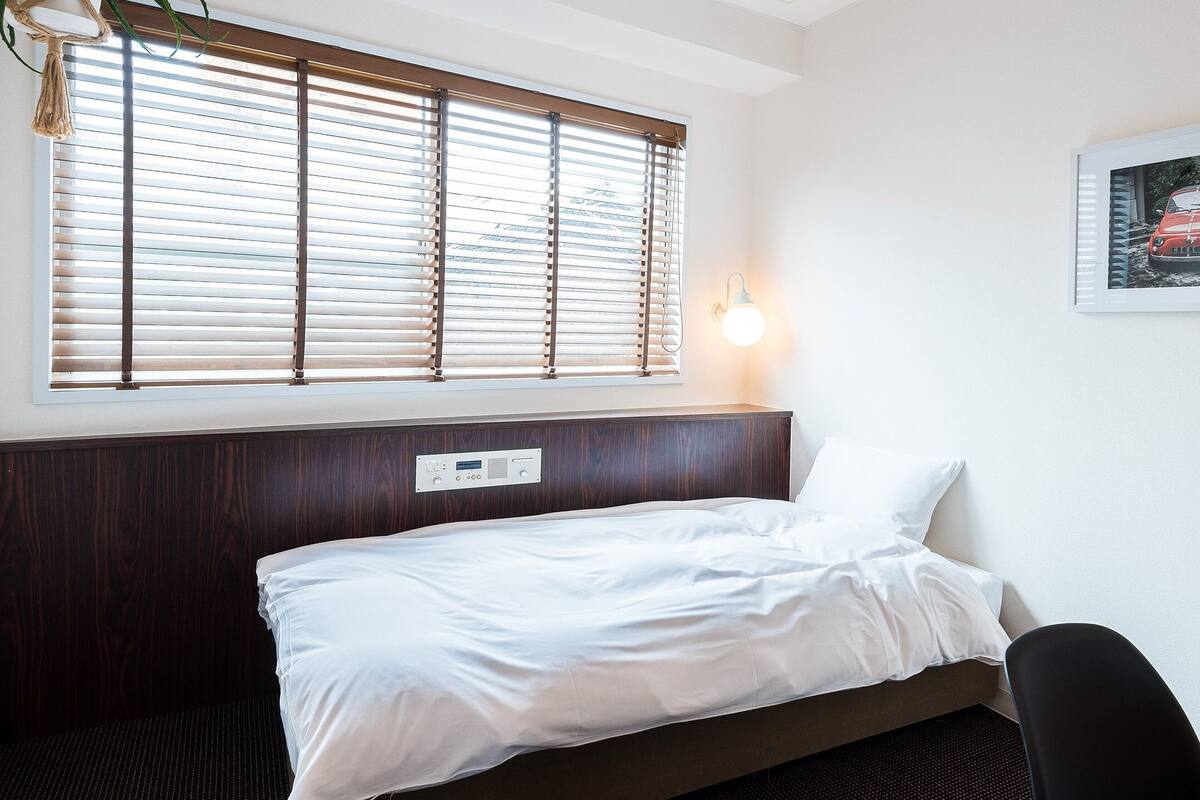 A hotel room with a bed, a headboard, a wall-mounted light, a window with blinds, and a framed picture on the wall.