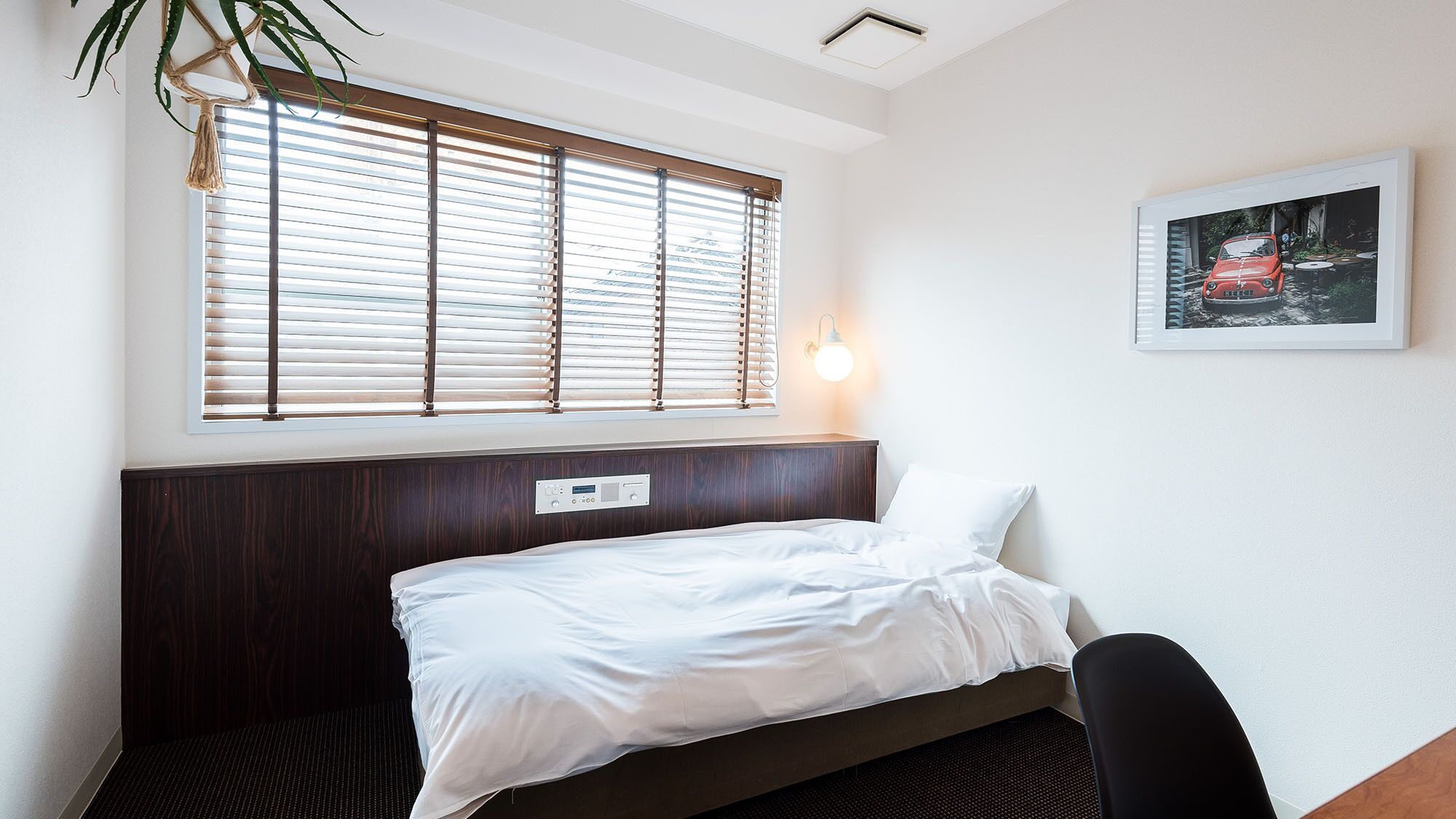 Une chambre d’hôtel avec un lit, une fenêtre avec des stores, une lampe fixée au mur et un tableau encadré.