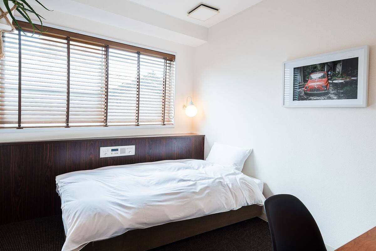 A hotel room with a bed, a window with blinds, a wall-mounted light, and a framed picture.