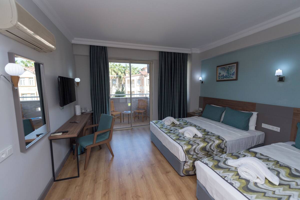 Quarto de hotel com escrivaninha, duas camas, uma cadeira, televisão e varanda com cortinas.