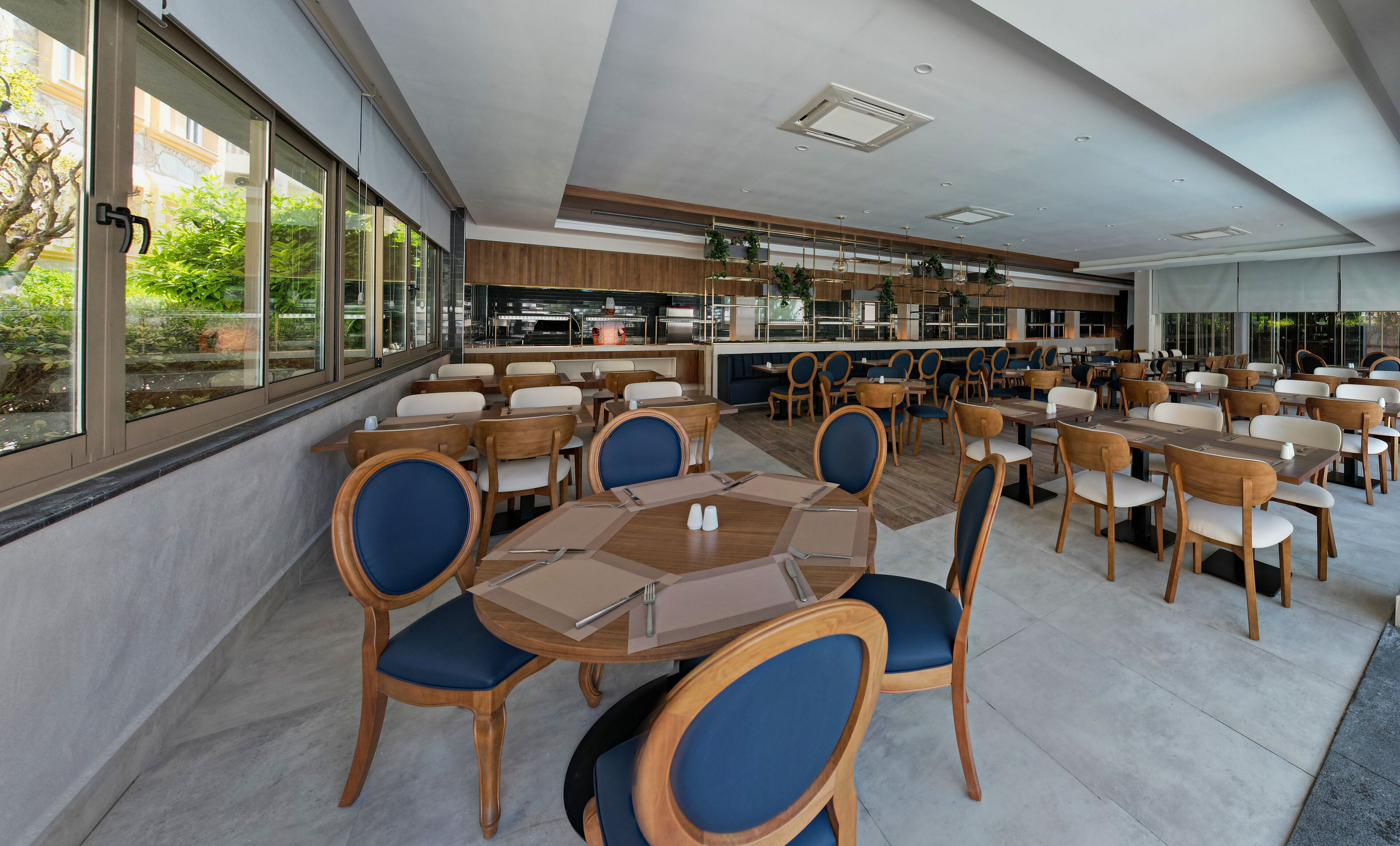 En moderne restaurant med træborde og -stole, store vinduer og et køkkenområde, der kan ses igennem glasset.