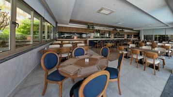 En moderne restaurant med træborde og -stole, store vinduer og et køkkenområde, der kan ses igennem glasset.