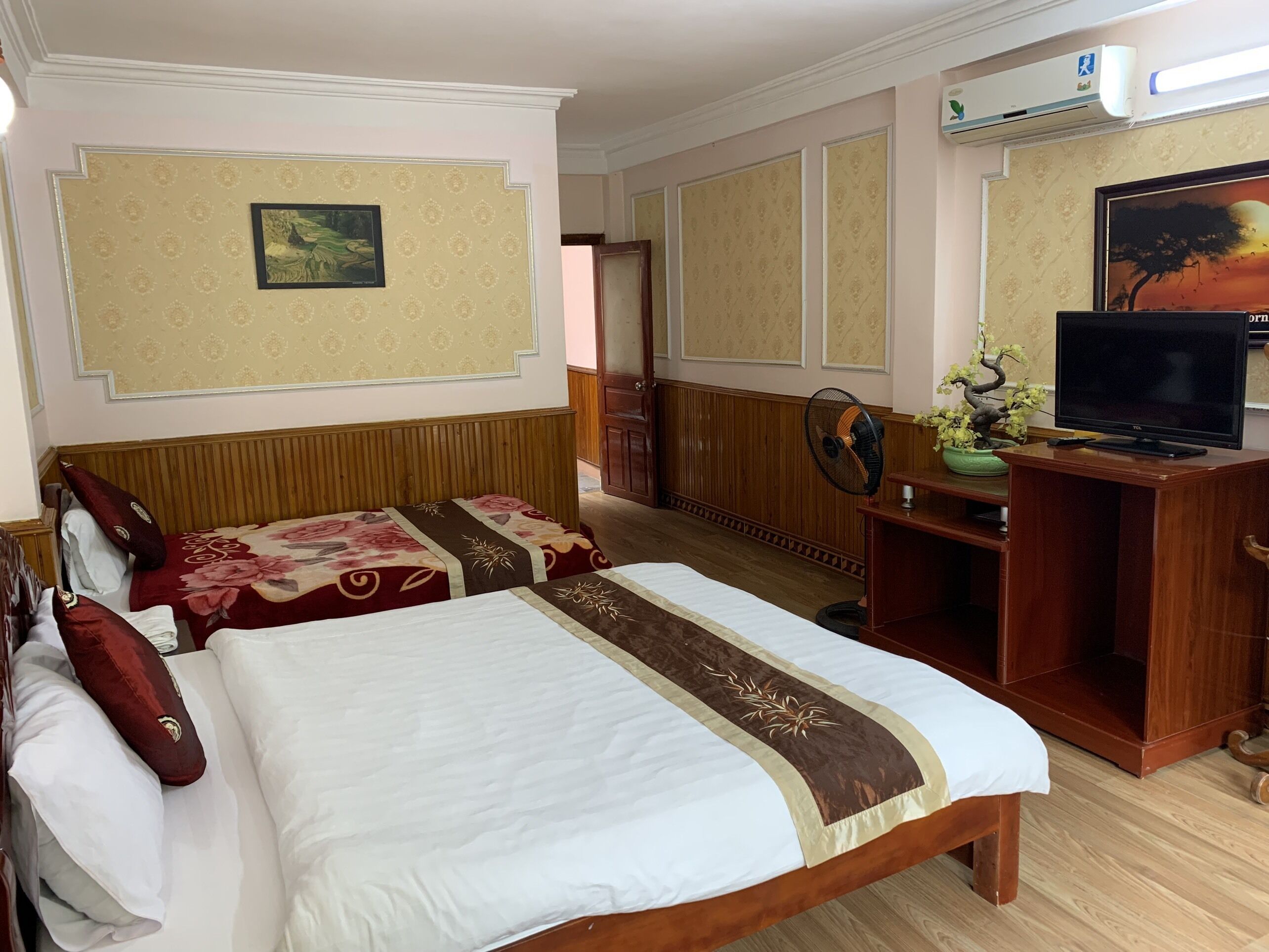 A hotel room with two beds, a TV, a fan, and a painting on the wall.