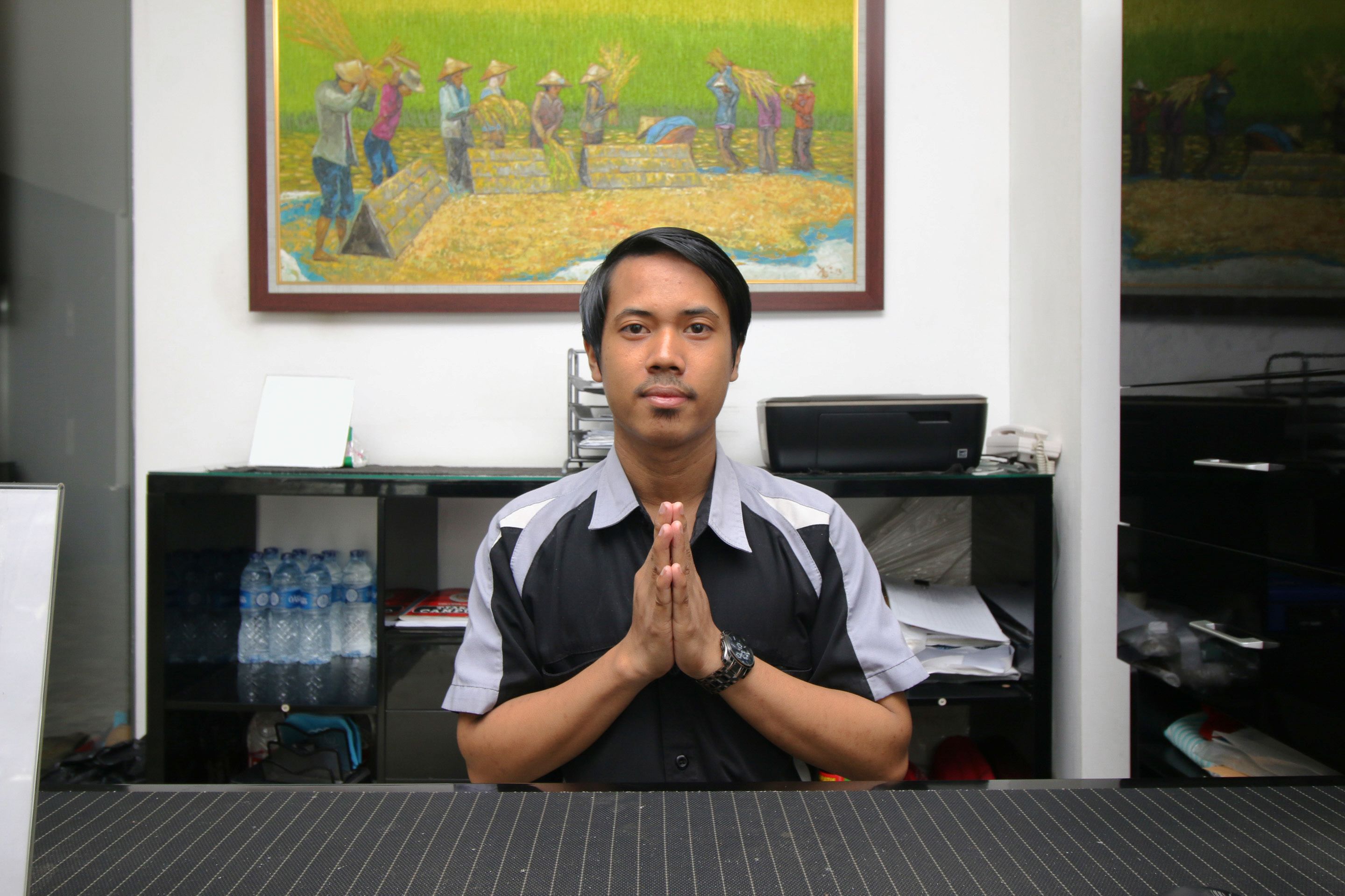 A person standing in an office with a desk, computer, and a painting of workers on the wall.