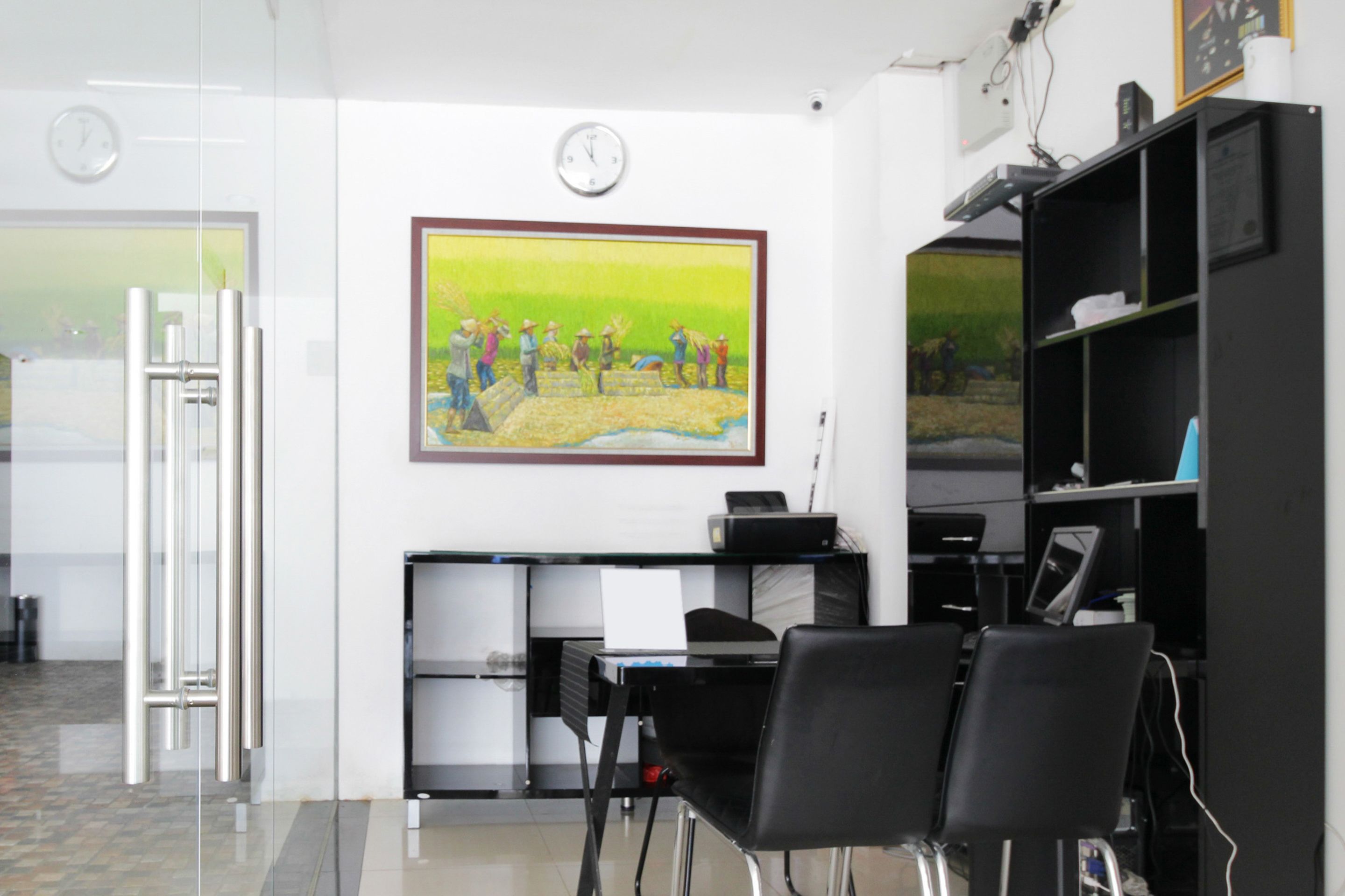 A modern office with a glass door, a desk with a computer, black chairs, and a painting on the wall.