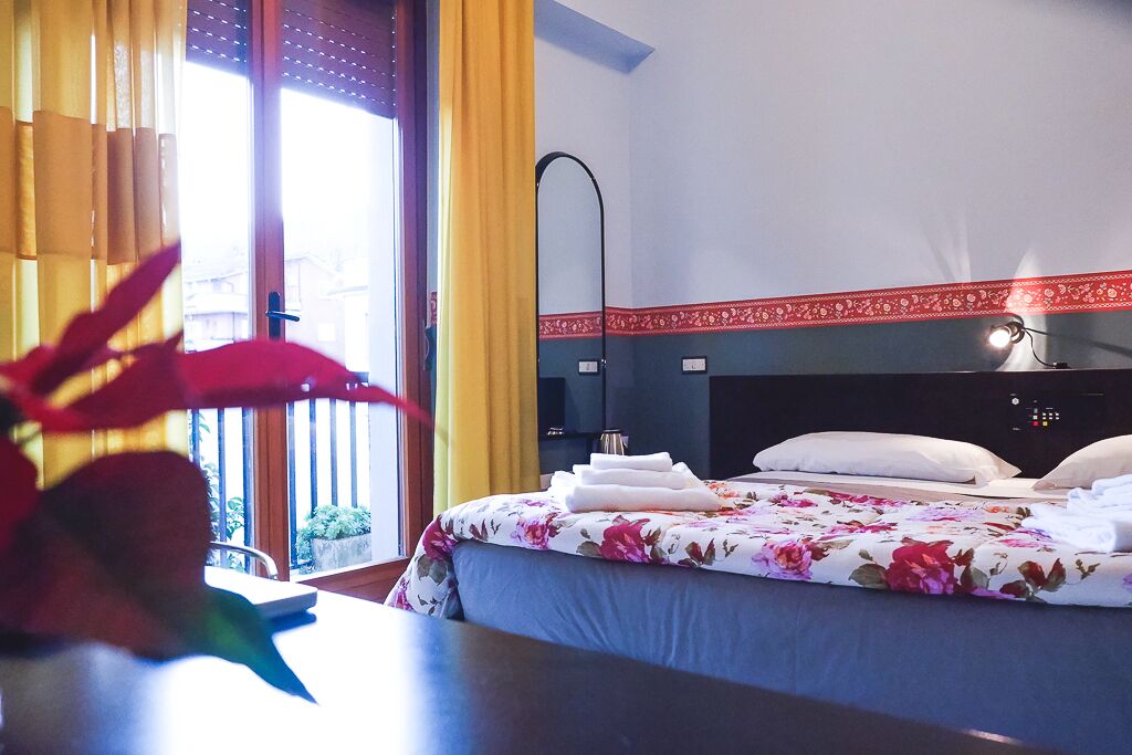 A hotel room with a bed, a bedside table, a window with curtains, and a view of the outside.
