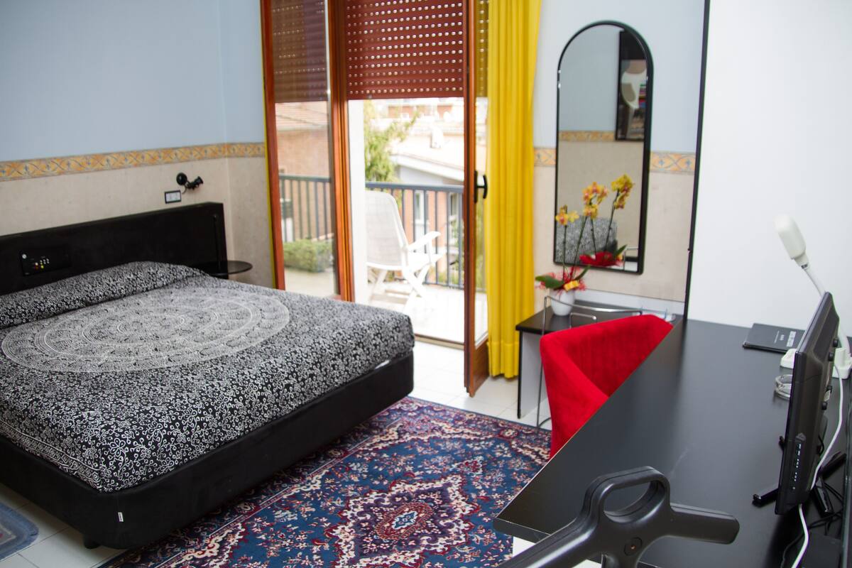 Une chambre à coucher comprenant un lit, un bureau, une chaise, un miroir et donnant sur l’extérieur.