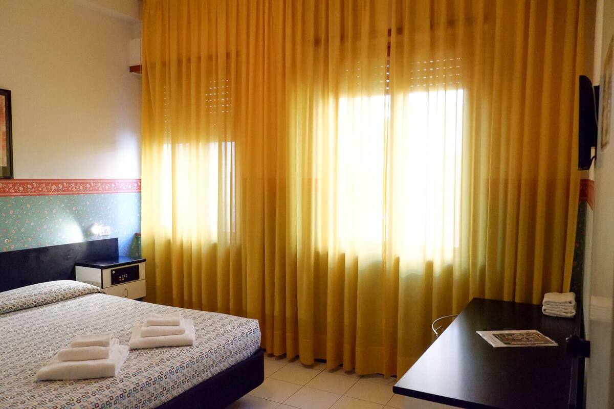 Une chambre d’hôtel avec un lit, un bureau et une fenêtre avec des rideaux jaunes.