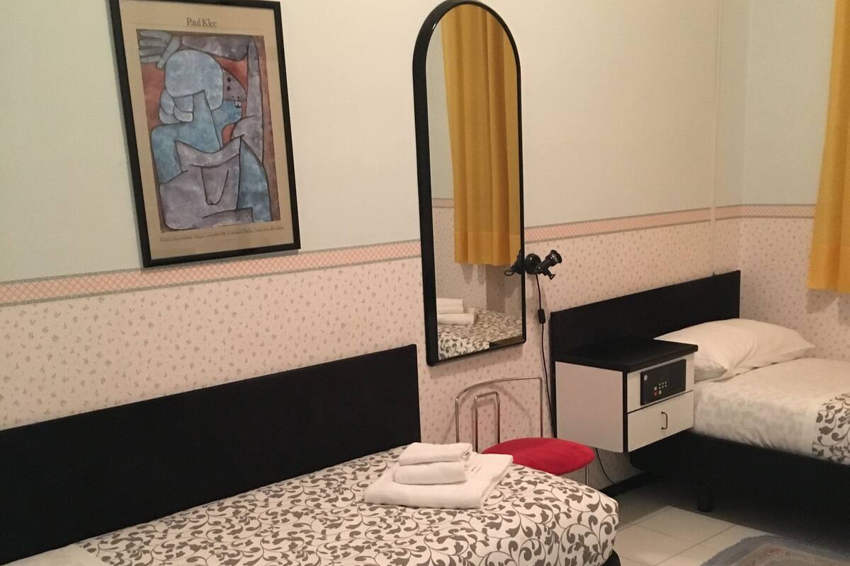 Une chambre d’hôtel avec deux lits simples, une petite table, un miroir, un tableau encadré et une fenêtre avec des rideaux.