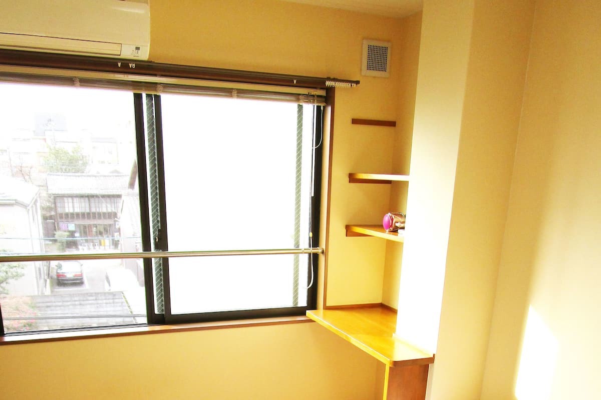 A room with tatami flooring, a bed with white bedding, a window with a view of the street, and a shelf with a pink object.