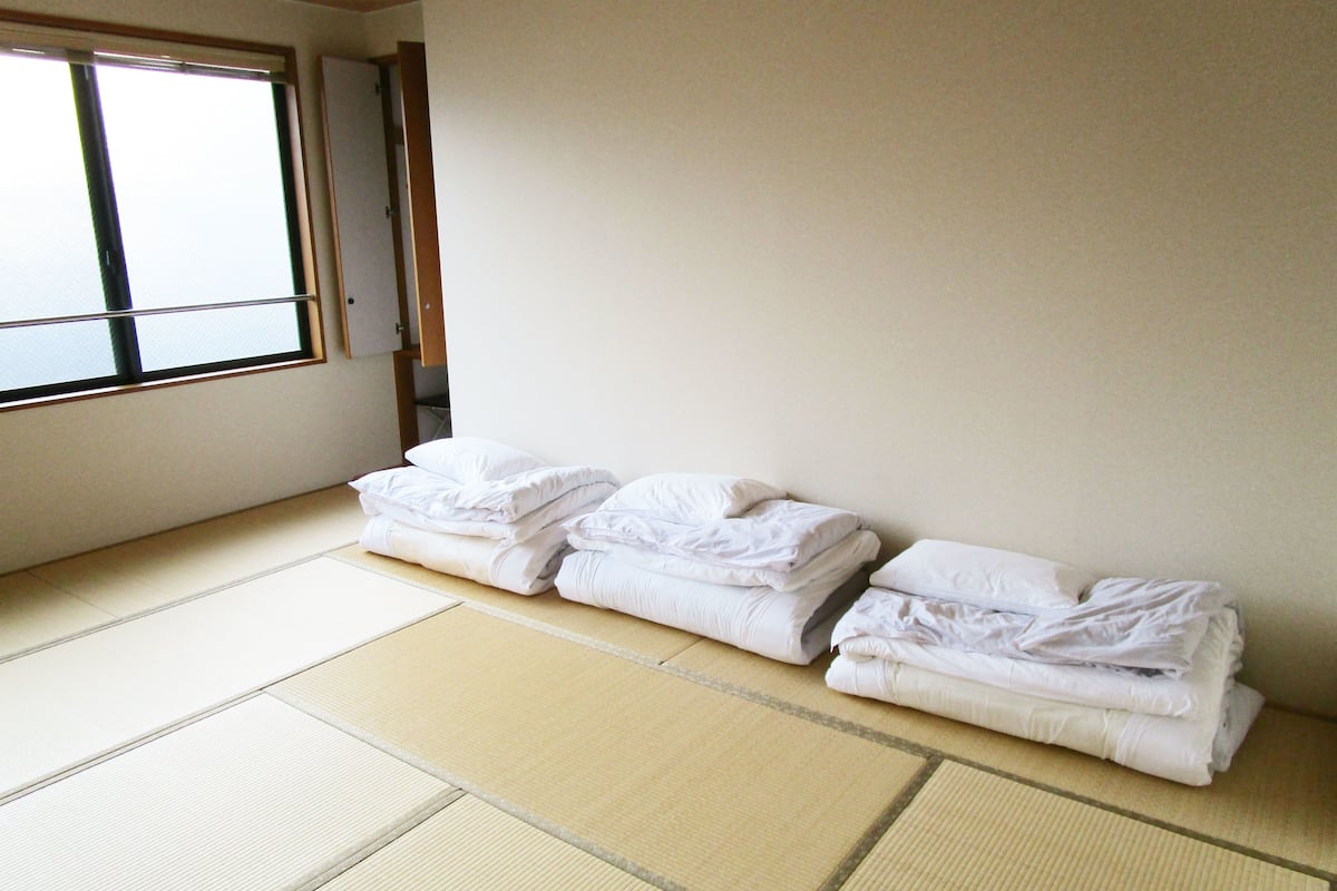 A room with tatami mats, a window, and neatly stacked white bedding.