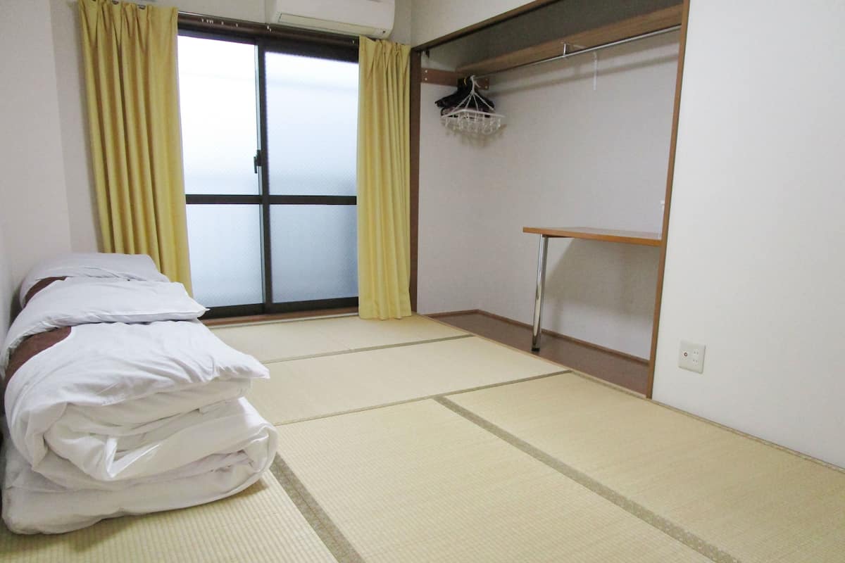 A room with tatami mat flooring, a bed with white bedding, a sliding door with yellow curtains, and a wooden shelf with a hanging rack.