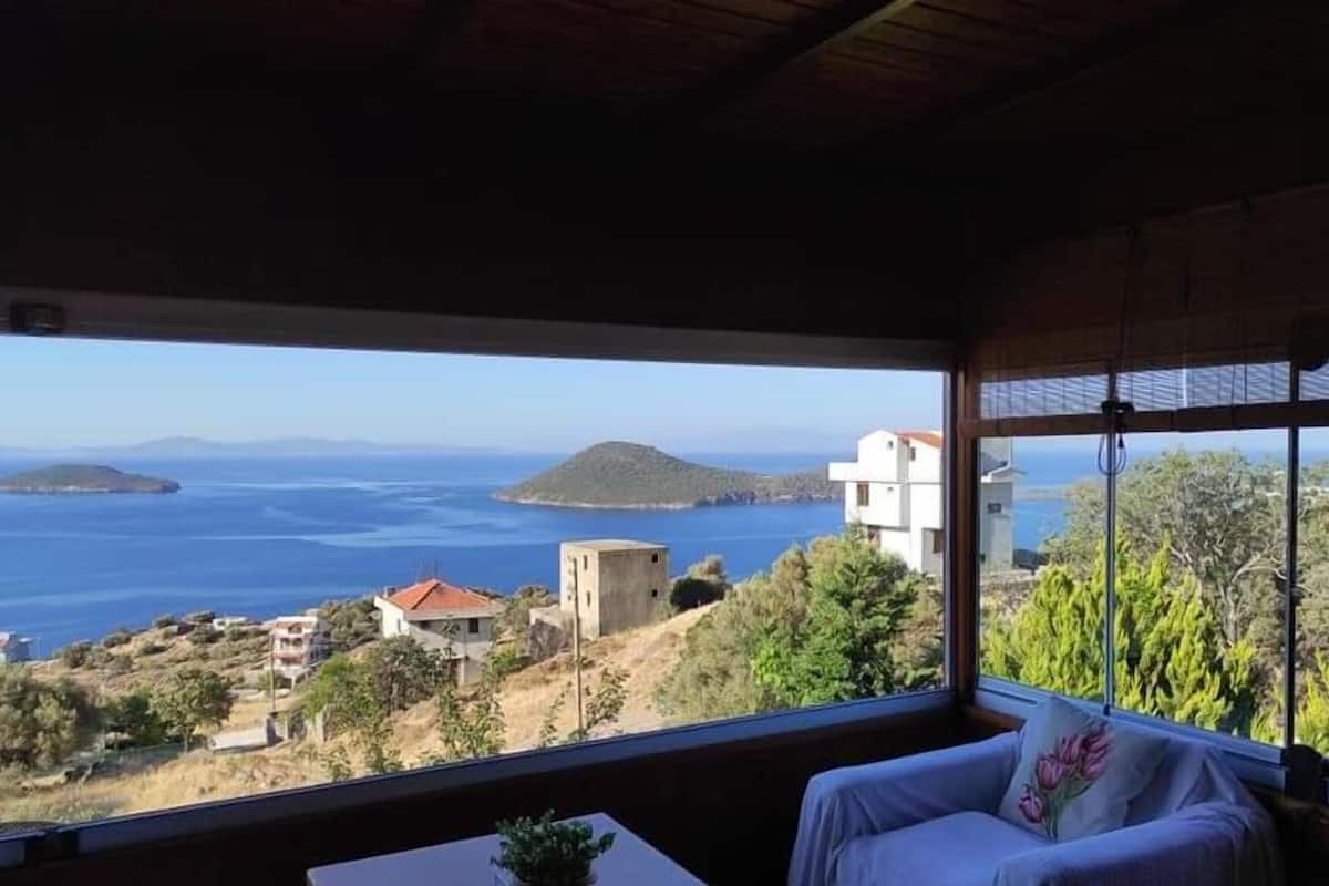 A room with a large window offering a view of the sea and hills.