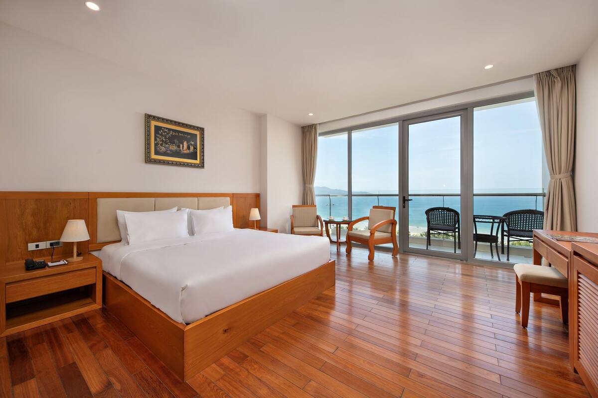 Une chambre d’hôtel dotée d’un grand lit, de tables de chevet en bois, d’un bureau avec des chaises et d’un balcon offrant une vue sur la mer.