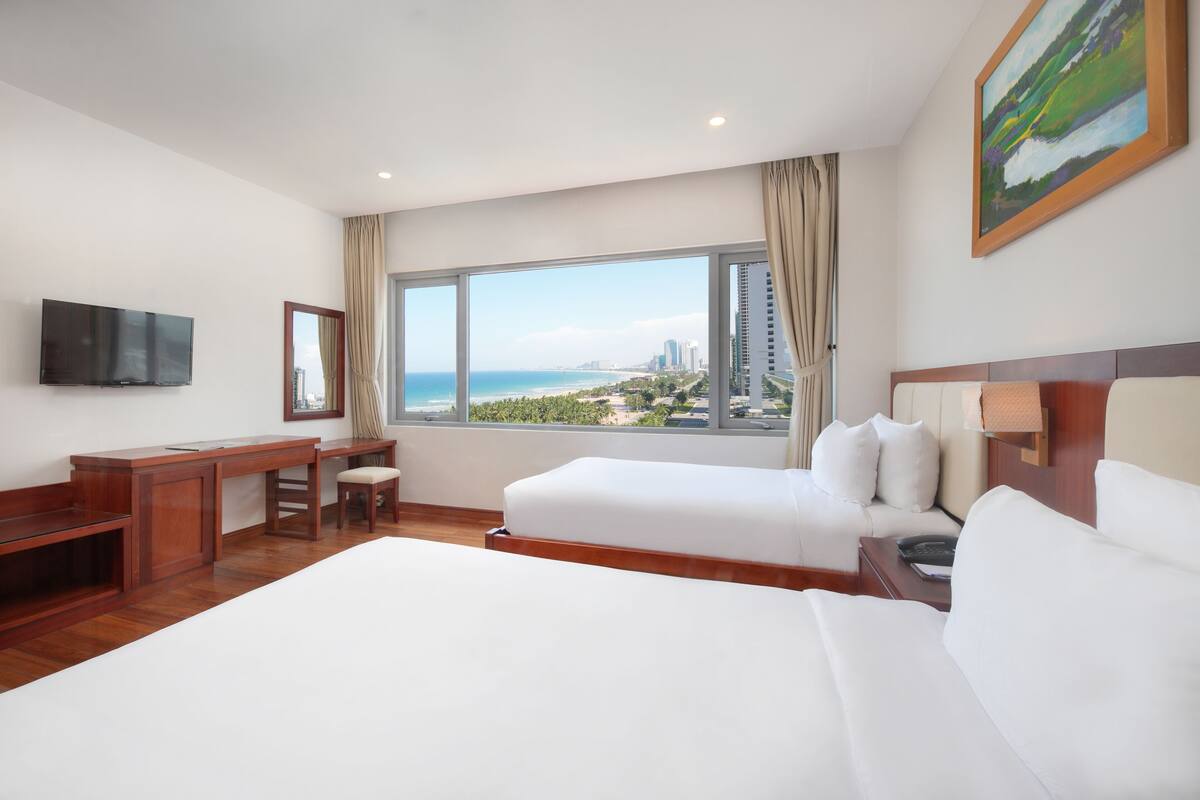 Une chambre d’hôtel avec un grand lit, un bureau, une chaise, une télévision et une fenêtre donnant sur la ville et l’océan.