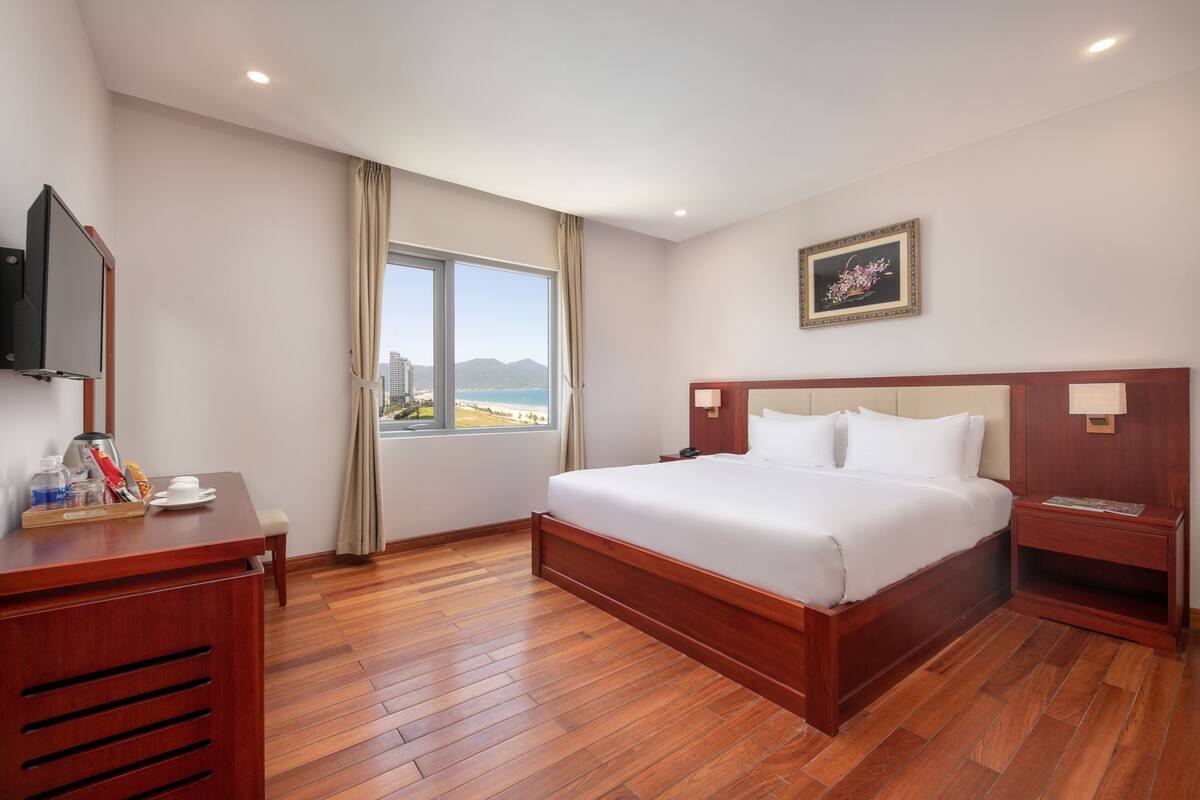 Une chambre d’hôtel avec un plancher en bois, un grand lit avec du linge de lit blanc, un bureau avec un plateau de petit-déjeuner, une télévision et une fenêtre donnant sur la mer et les montagnes.
