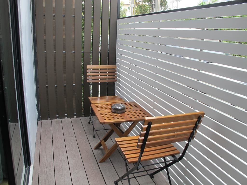 Uma varanda com uma mesa de madeira e duas cadeiras, grade de metal cinzenta e vista para o verde.
