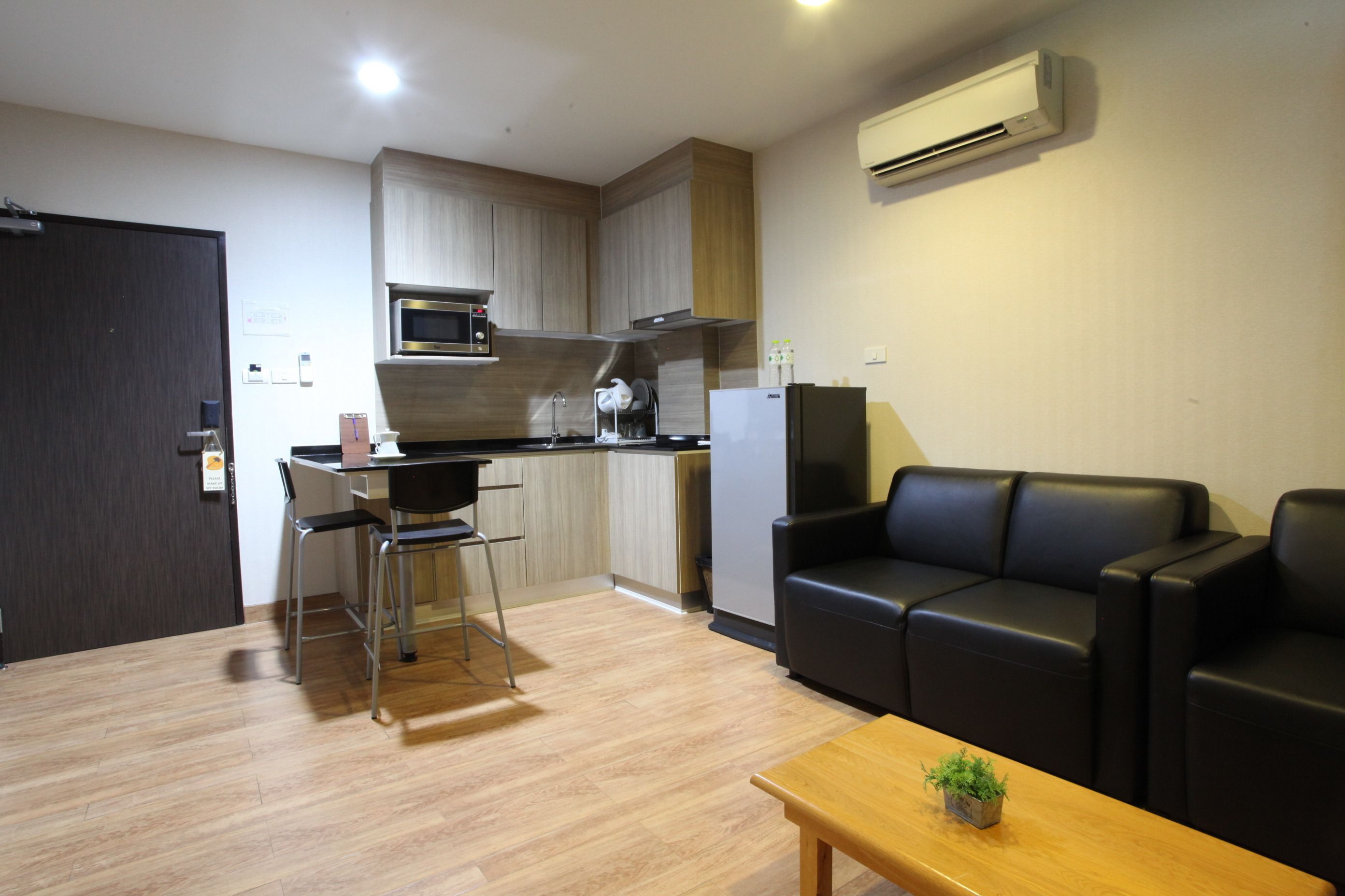 Un espacio habitable compacto con kitchenette, un sofá de cuero negro, una mesa de centro de madera y una planta pequeña.