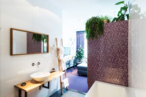 A modern bathroom with a white sink, wooden bench, and a large mirror. There is a shower area with a purple tiled wall and a hanging plant. - Wolf Hotel Kitchen & Bar (Alkmaar)