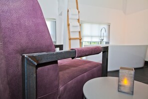 A purple upholstered chair with a wooden armrest and a small table with a candle. A wooden ladder and a white bathtub are visible in the background. - Wolf Hotel Kitchen & Bar (Alkmaar)
