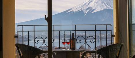 A balcony with a view of a snow-capped mountain, a table with two wine glasses and a bottle, and wicker chairs.