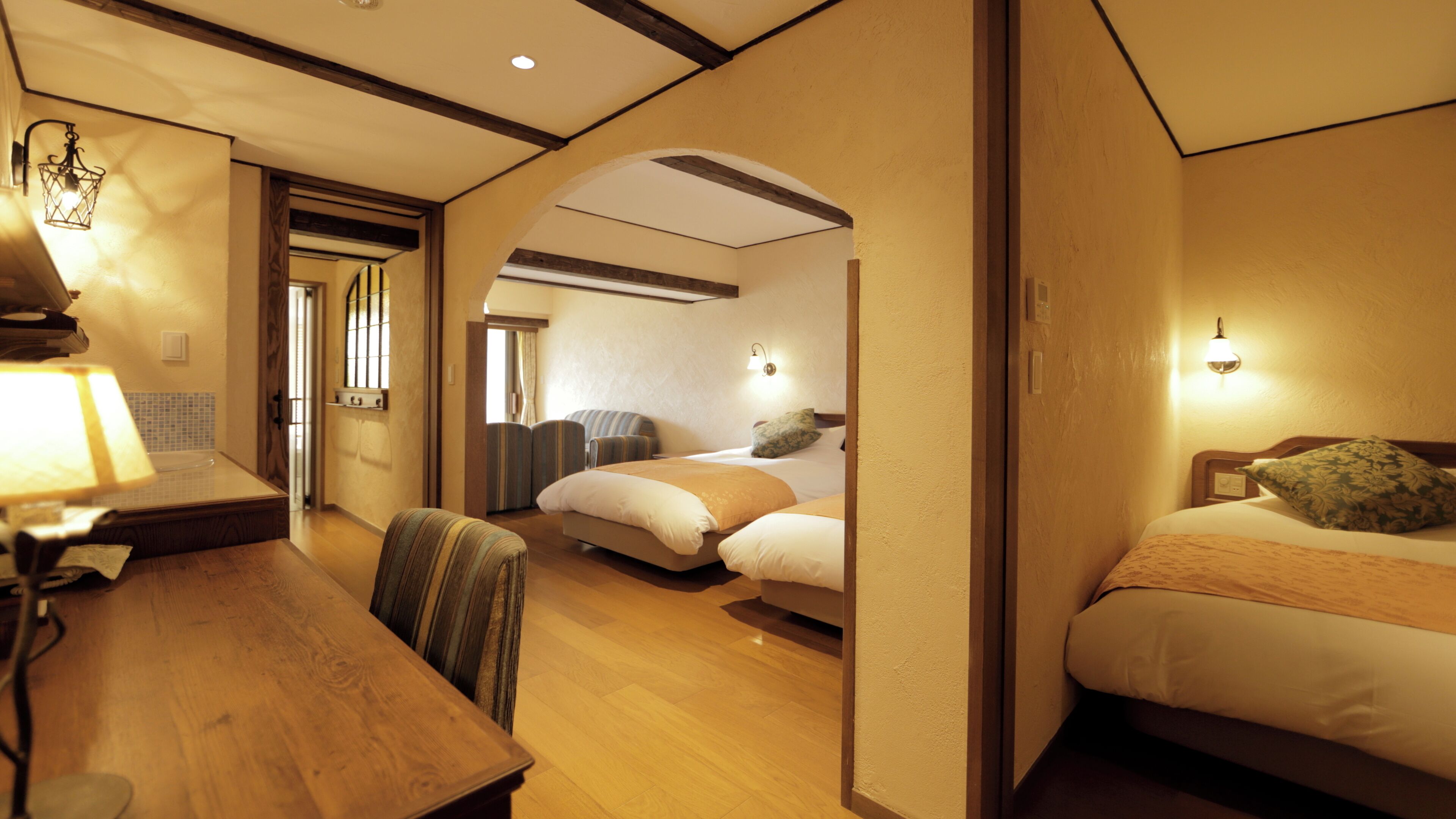 Quarto de hotel com escrivaninha de madeira, duas camas, área de estar com uma cadeira e uma mesinha.