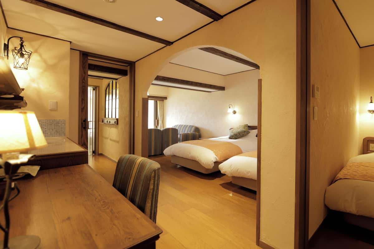 Quarto de hotel com escrivaninha de madeira, duas camas, área de estar com uma cadeira e uma mesinha.
