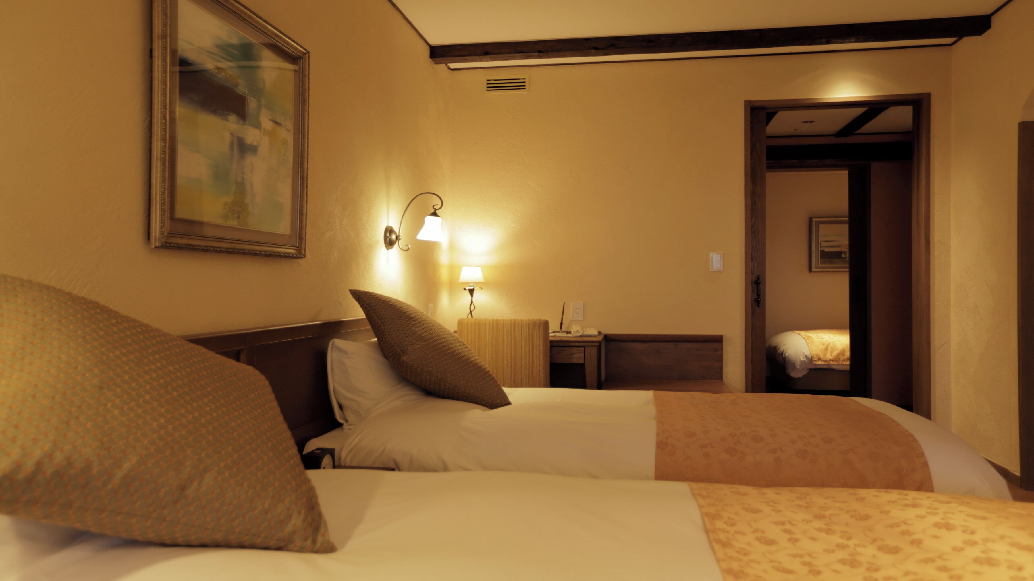 Quarto de hotel com duas camas, um quadro na parede, uma mesa de cabeceira com abajur e uma porta que dá para outro quarto.
