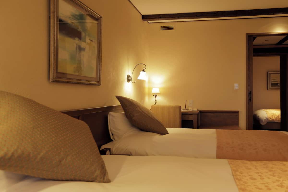 Quarto de hotel com duas camas, um quadro na parede, uma mesa de cabeceira com abajur e uma porta que dá para outro quarto.