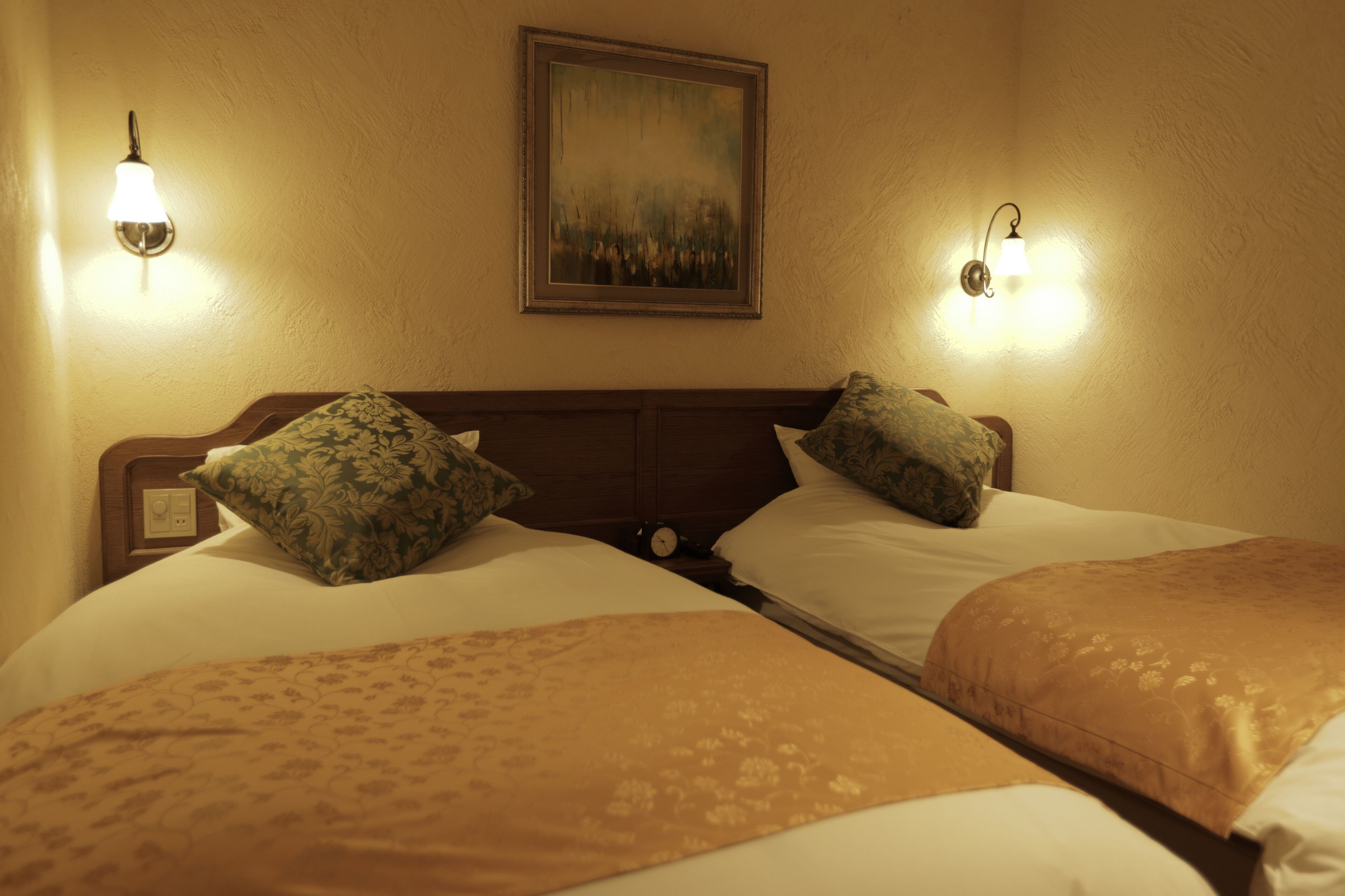 Een hotelkamer met twee bedden, elk voorzien van beddengoed en kussens met bloemenprint. Aan weerszijden van een schilderij boven de bedden hangen wandlampen.