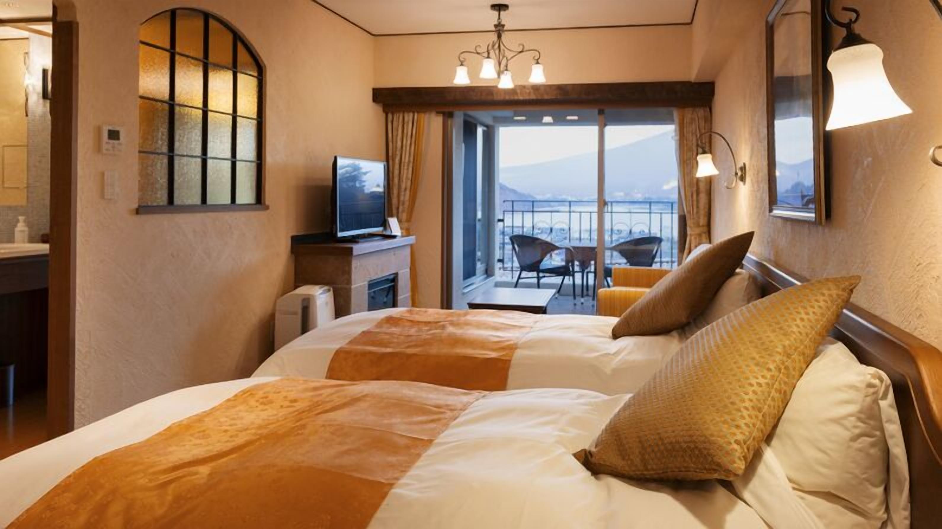 Quarto de hotel com cama, televisão, varanda com cadeiras e vista para as montanhas.