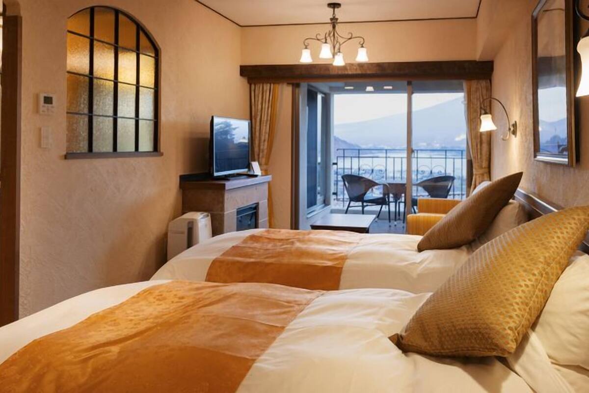 Quarto de hotel com cama, televisão, varanda com cadeiras e vista para as montanhas.