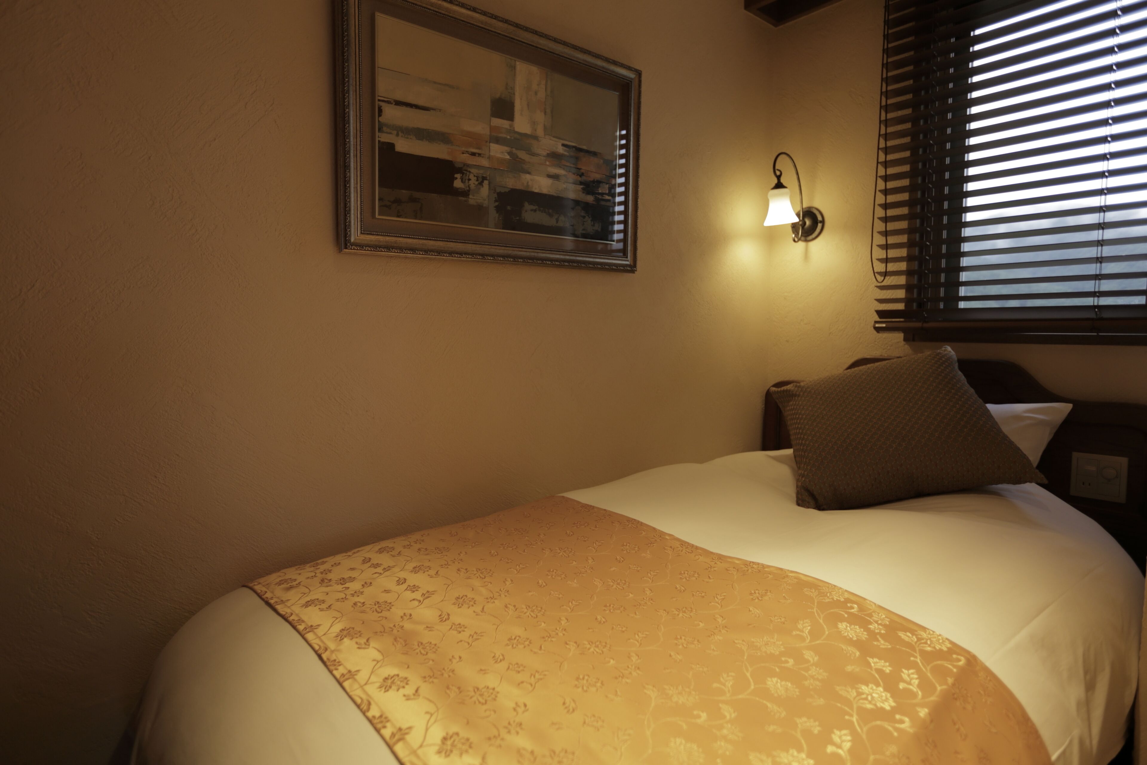 Una cama bien tendida con una manta de estampado floral, una obra de arte enmarcada en la pared y una lámpara fijada a la pared.