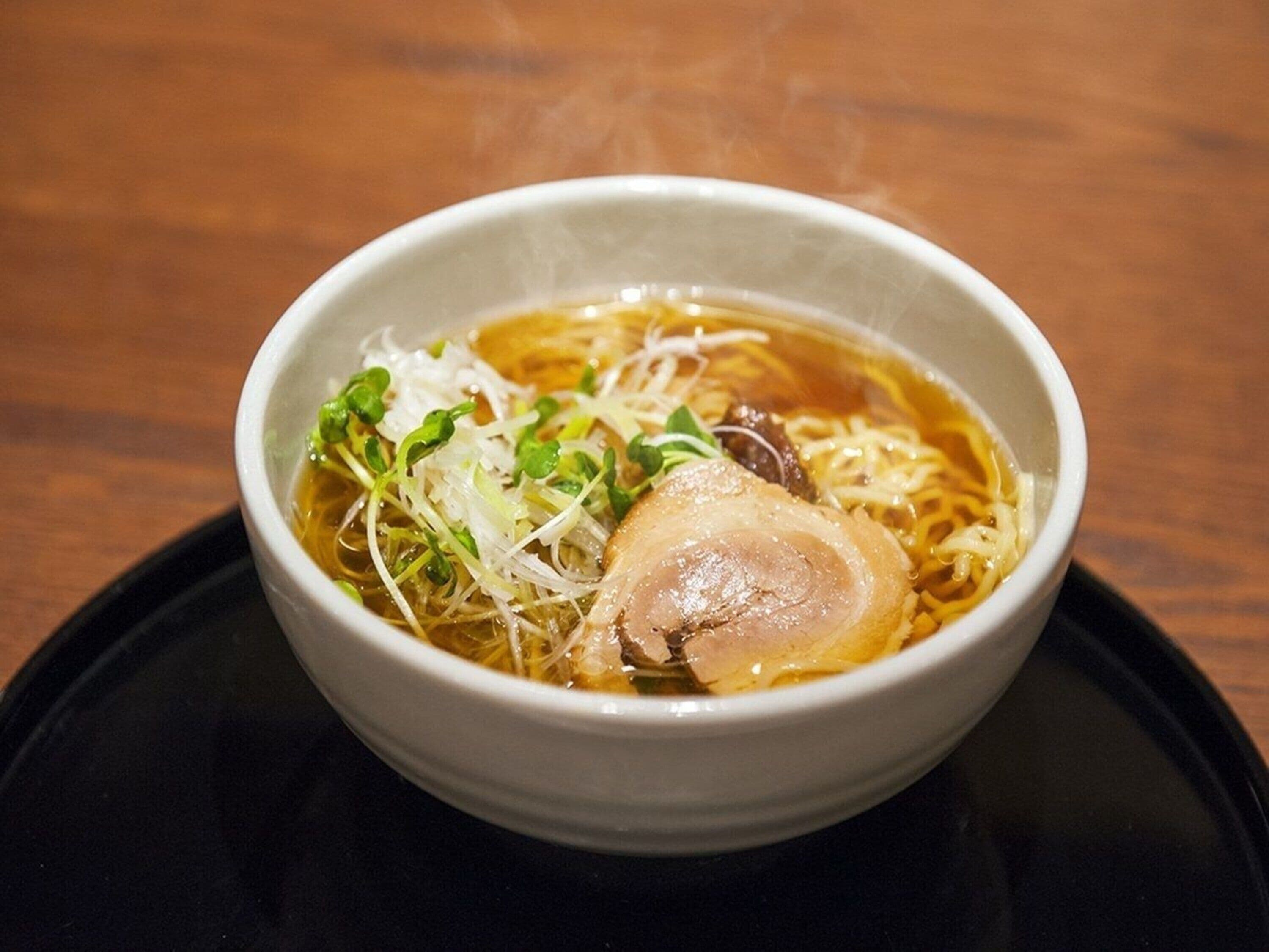 A bowl of noodle soup with meat and greens on a black plate.
