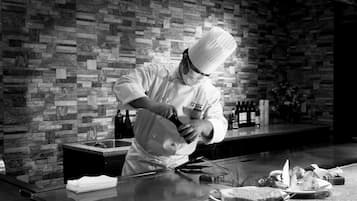 A chef in a professional kitchen with a brick wall background, a stainless steel countertop, and various kitchen utensils and bottles.