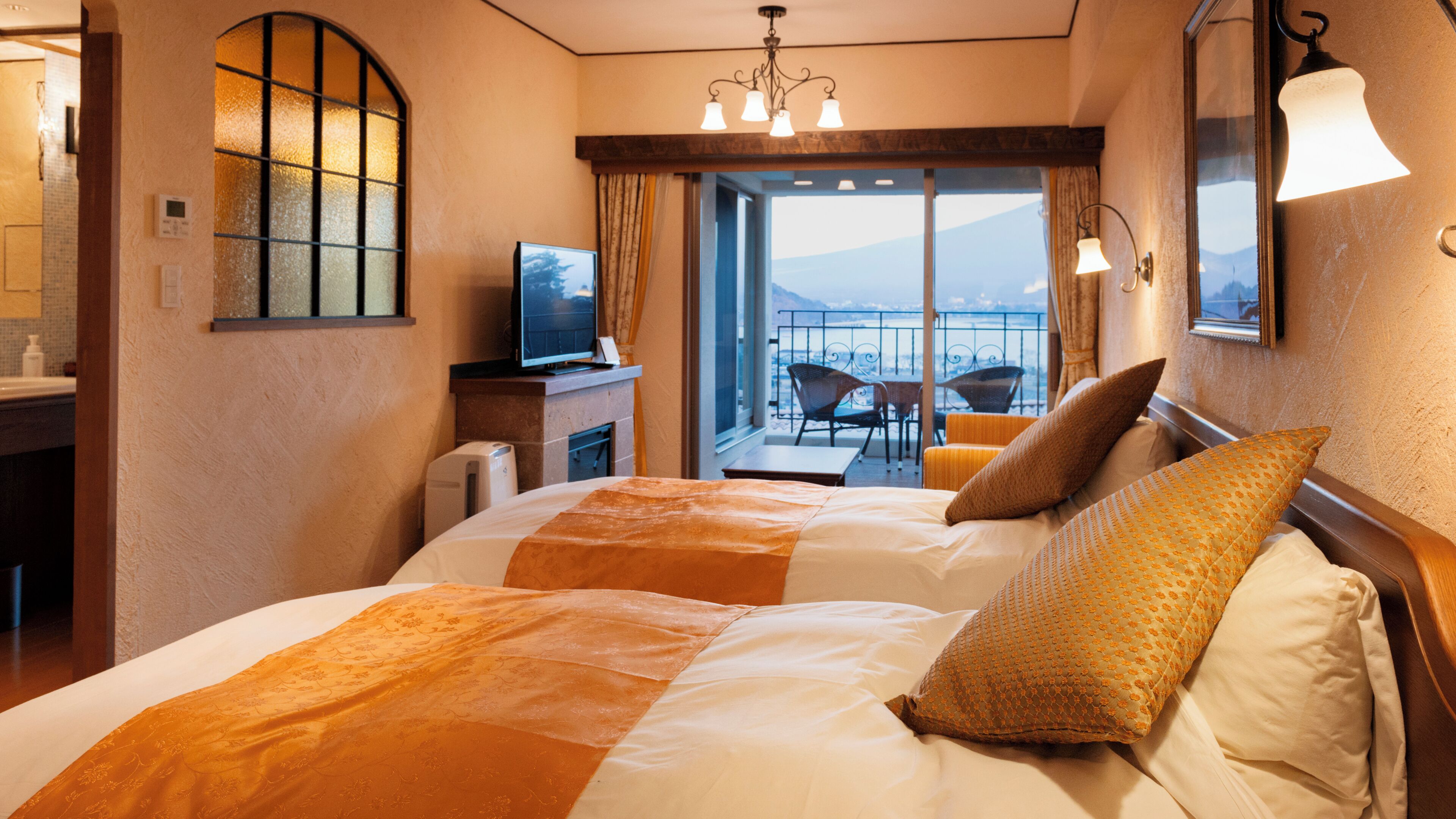 Quarto de hotel com cama, televisão, varanda com cadeira e vista para as montanhas.