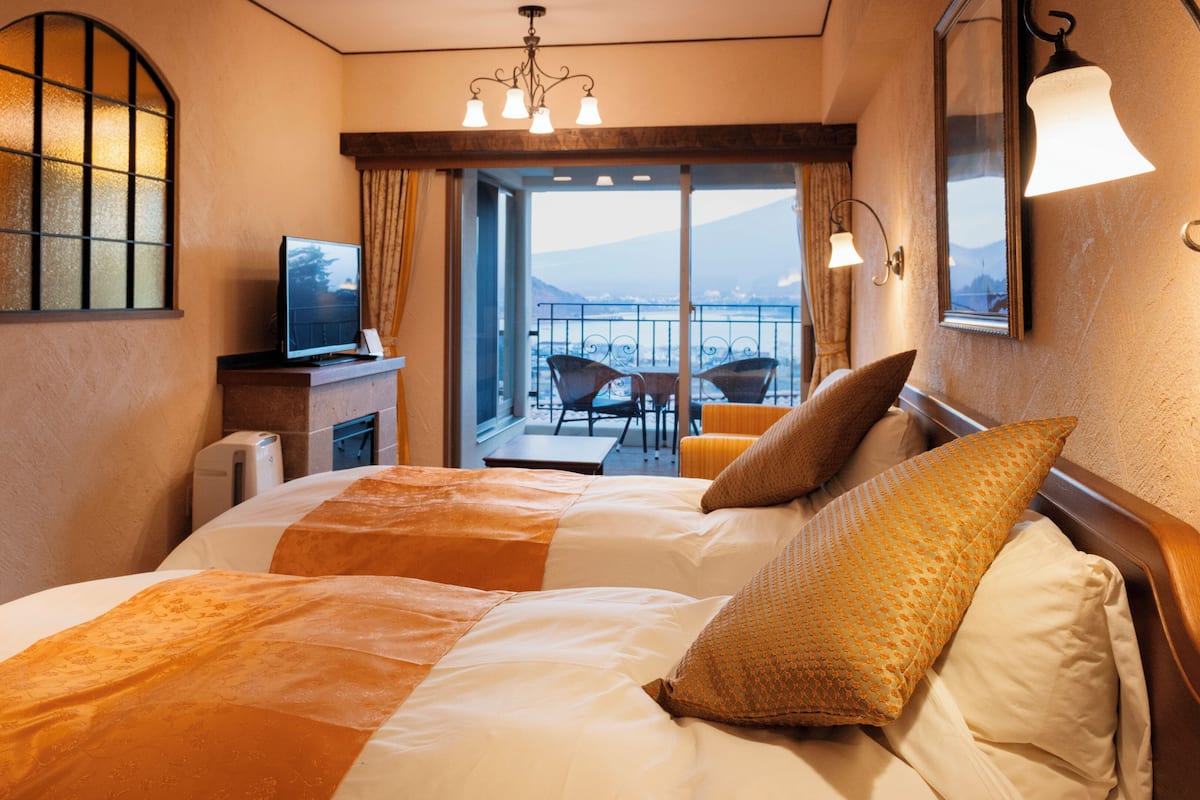 Quarto de hotel com cama, televisão, varanda com cadeira e vista para as montanhas.