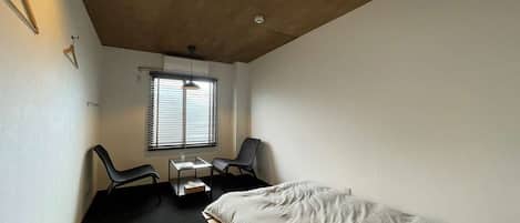 A small, modern bedroom with a single bed, a desk with a chair, and a window with blinds.