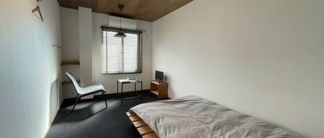 A bedroom with a single bed, a desk with a chair, a window with blinds, and a hanging light fixture.
