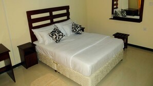 A neatly made bed with white bedding and patterned pillows, a wooden headboard, a bedside table, and a nightstand. A mirror hangs on the wall above the bed. - Dilray Villa (Harispattuwa)