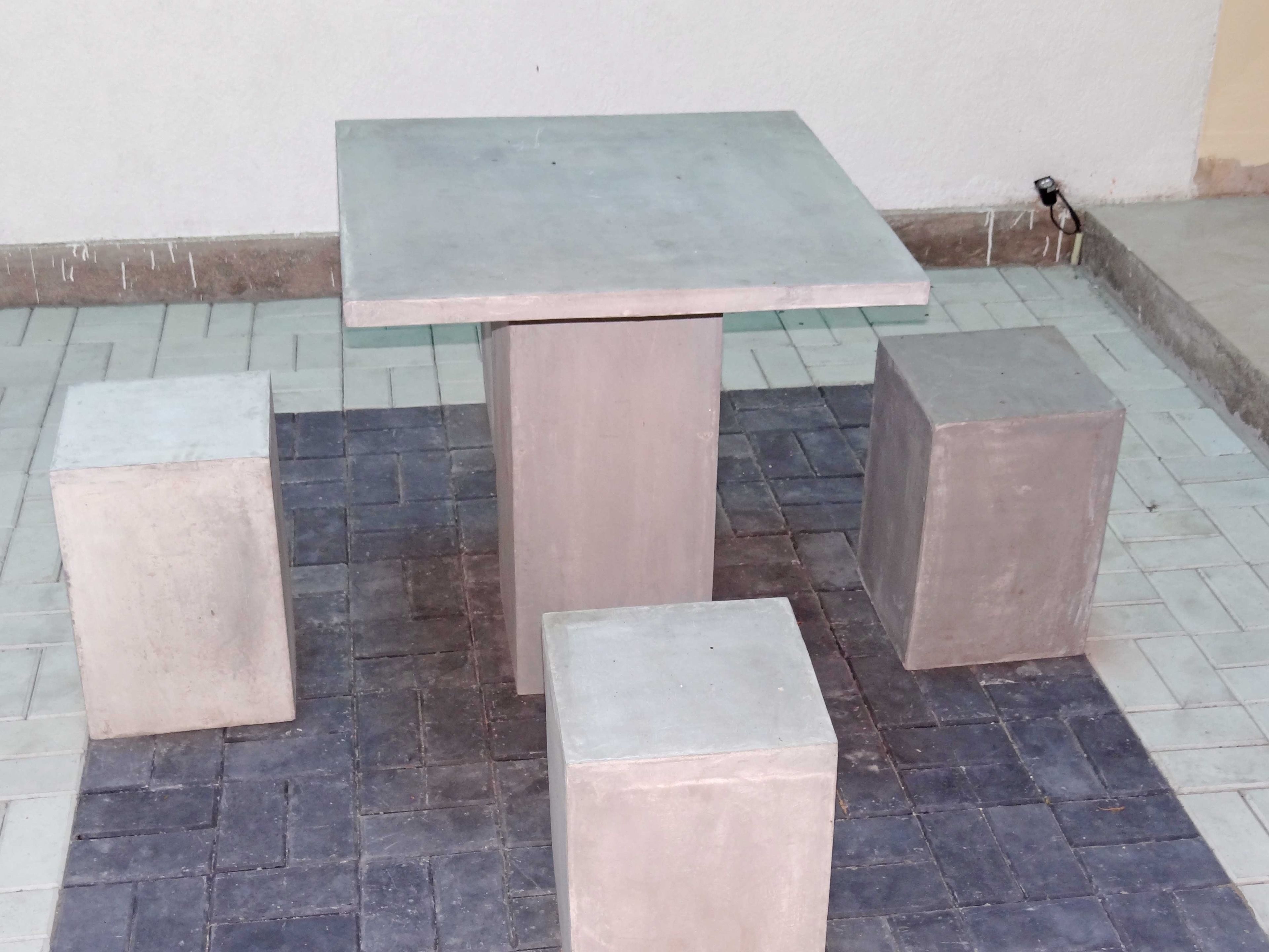Una mesa cuadrada con tapa rectangular y base cilíndrica, rodeada por cuatro taburetes cuadrados.
