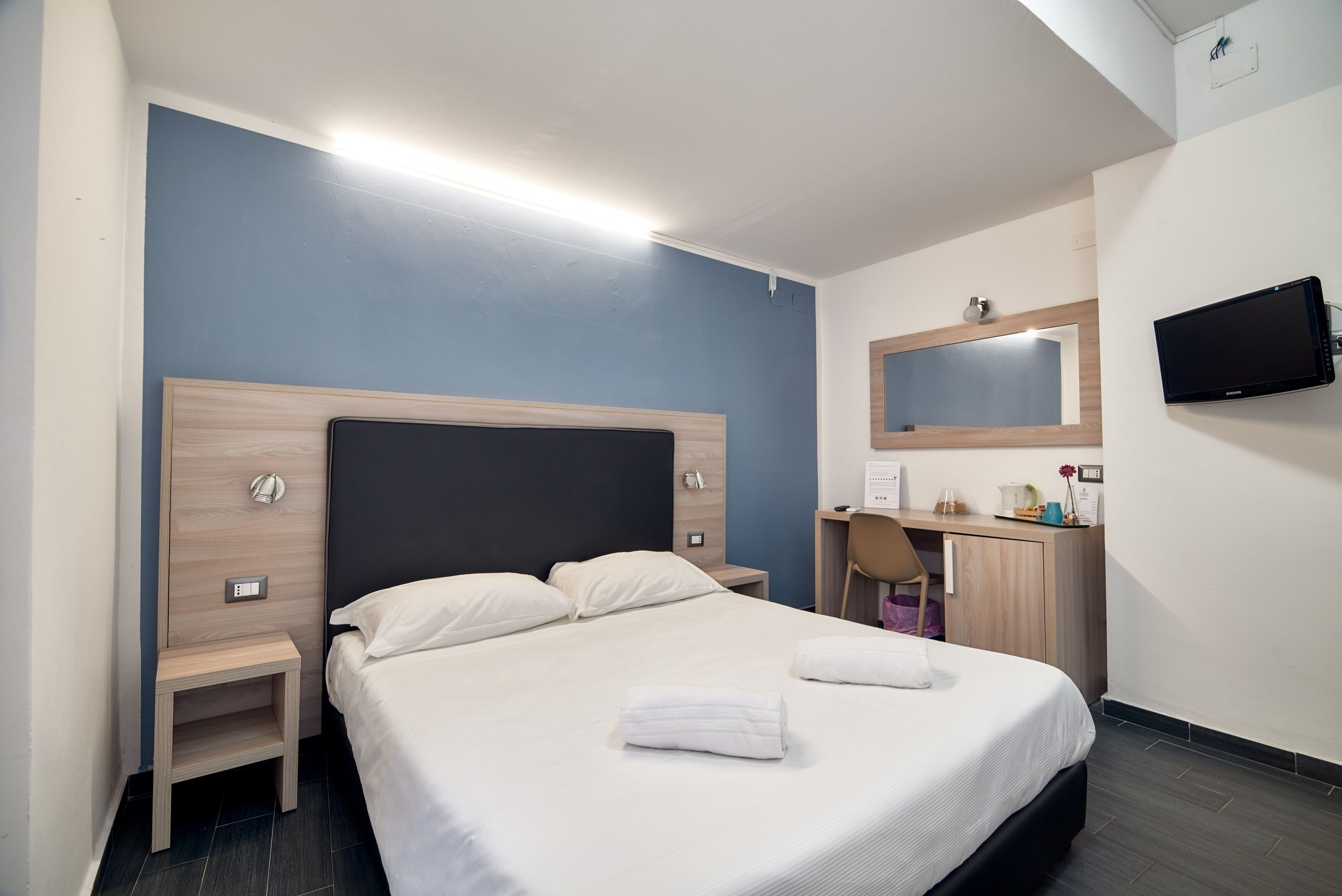A hotel room with a bed, bedside tables, a desk with a chair, a flat-screen TV, and a mirror on the wall.