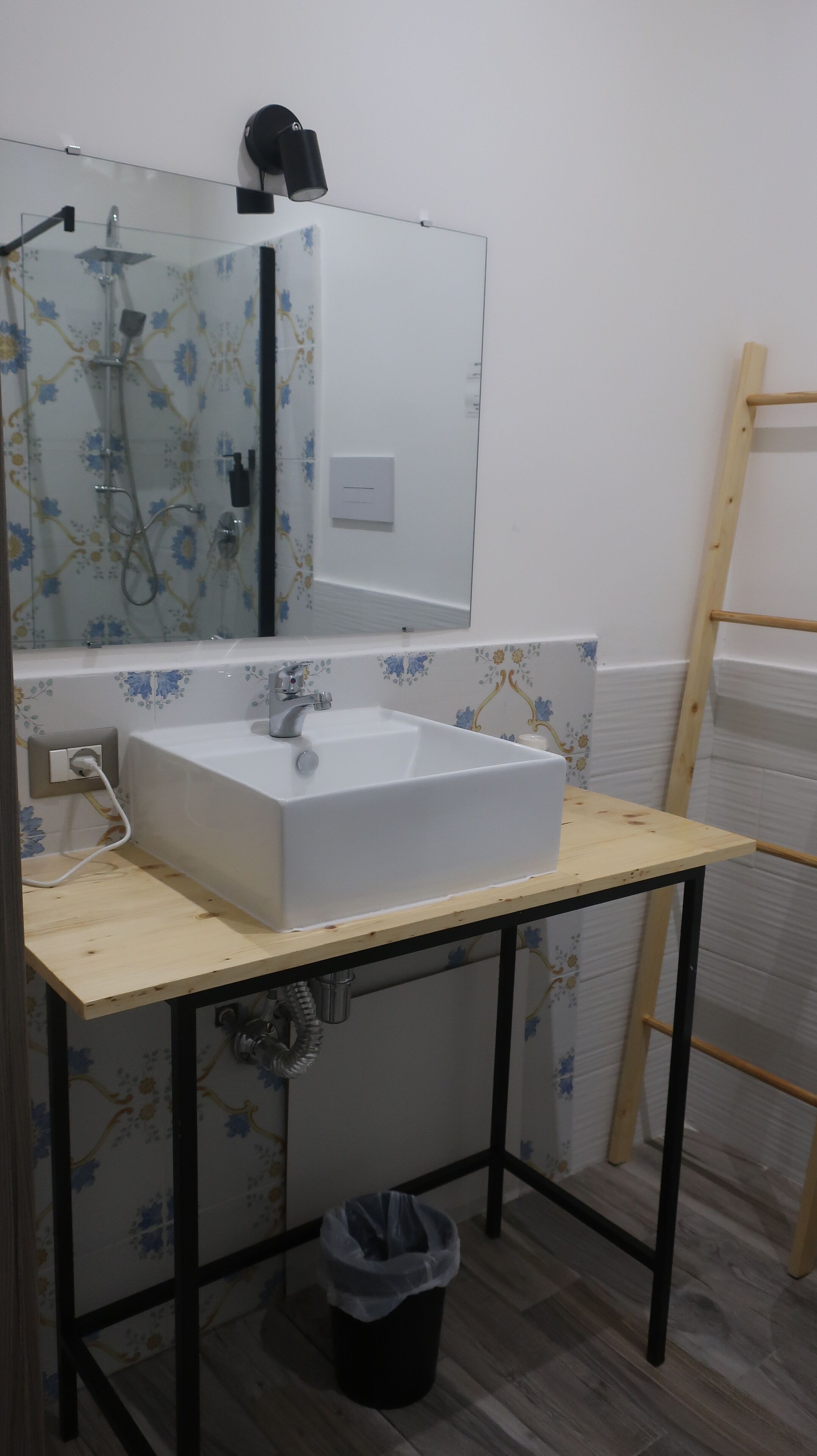 A bathroom with a white sink, a mirror, and a shower. There is a wooden stool and a ladder against the wall.