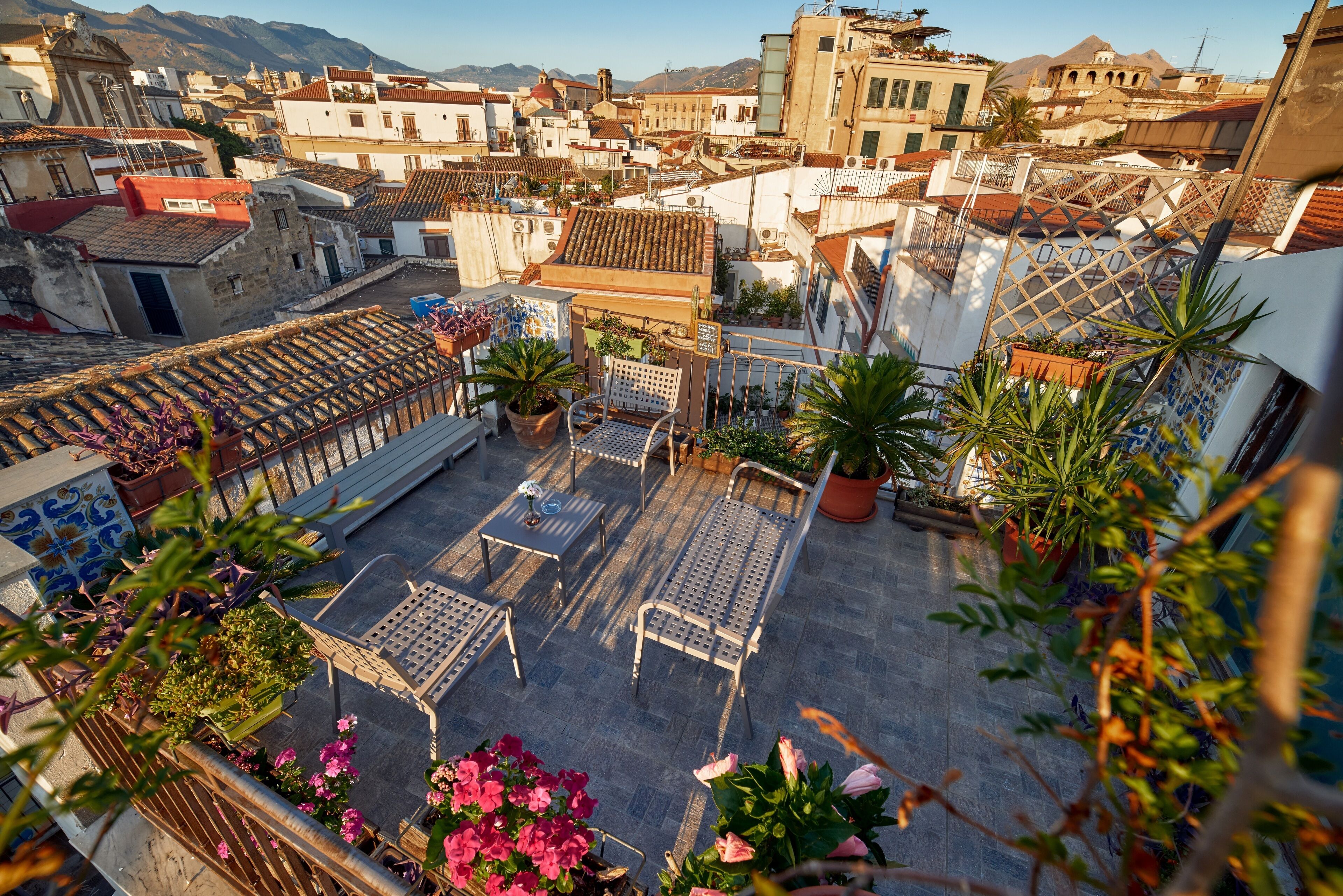 A rooftop terrace with potted plants, outdoor furniture, and a view of the surrounding buildings.